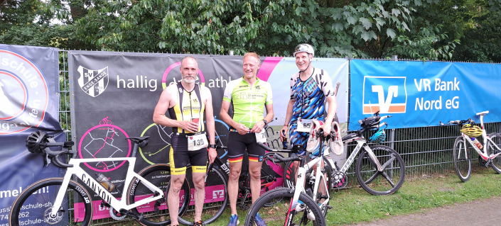 Celler Triathleten mit Fahrrädern vor dem Plakat des Hallig Dreeathlon 2025