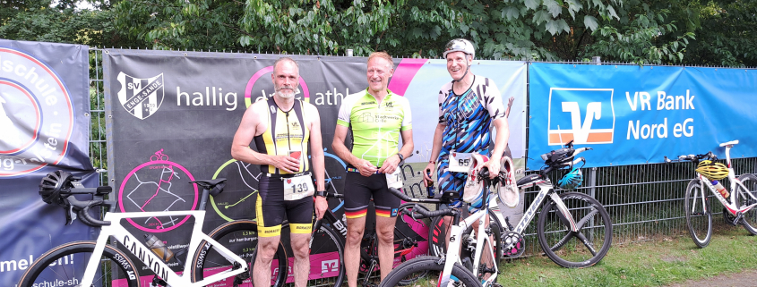 Celler Triathleten mit Fahrrädern vor dem Plakat des Hallig Dreeathlon 2025